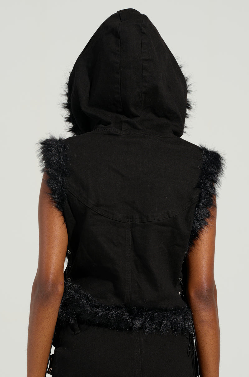 A person wearing the DELANEY FUR TRIM DENIM VEST, a sleeveless and hooded black vest with faux fur trim, stands facing away. The cropped silhouette and lace-up sides create a distinctive and stylish look against a neutral background.