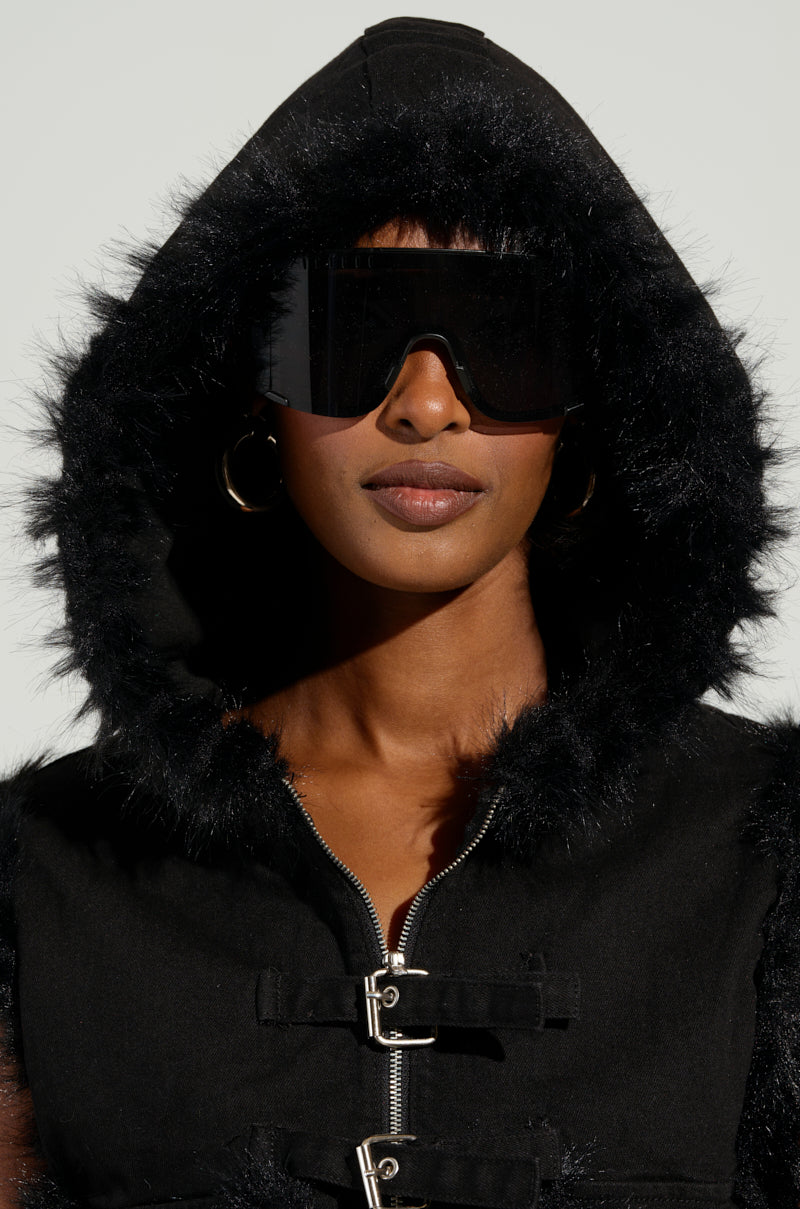 A person wearing the DELANEY FUR TRIM DENIM VEST, oversized sunglasses, and gold hoop earrings gazes directly at the camera against a plain background.