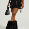 A person in the DELANEY FUR TRIM MINI SKIRT with metal eyelets, styled with matching black fur boots and a small black handbag, standing against a plain, light-colored background.