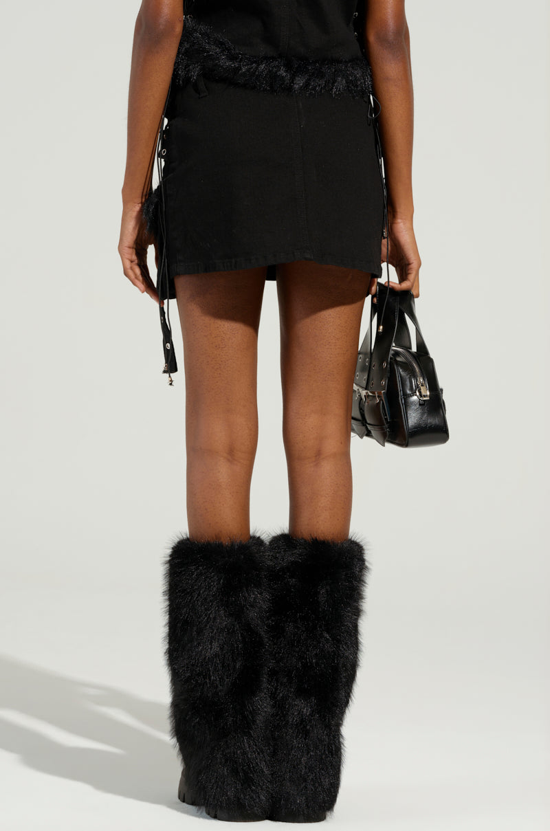 A person is standing with their back to the camera, wearing the DELANEY FUR TRIM MINI SKIRT and fuzzy black boots. They are holding a small black handbag in their right hand against a light gray background.