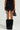 A person is standing with their back to the camera, wearing the DELANEY FUR TRIM MINI SKIRT and fuzzy black boots. They are holding a small black handbag in their right hand against a light gray background.