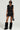 Wearing the DELANEY FUR TRIM MINI SKIRT with a black sleeveless outfit featuring fur accents, paired seamlessly with a studded belt and striking black fur boots, a person exudes confidence. Large sunglasses emphasize their look as they hold a small black handbag against the unadorned, light background.