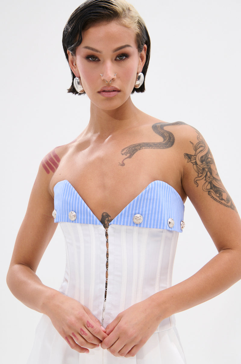 A person with short, dark hair and a blonde streak wears the strapless KLYA CORSET in white and blue with decorative buttons. Tattoos, facial piercings, and large hoop earrings add edge to their structured, seductive look.