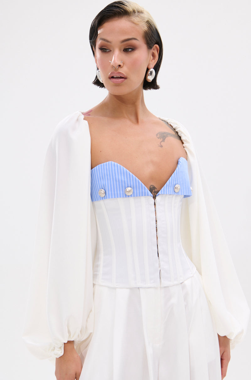 A person with short, dark hair and a blonde streak wears a white outfit with billowy sleeves and the KLYA CORSET, which features blue-and-white stripes, buttons, and nautical flair. Accessories include earrings, a nose ring, and tattoos.