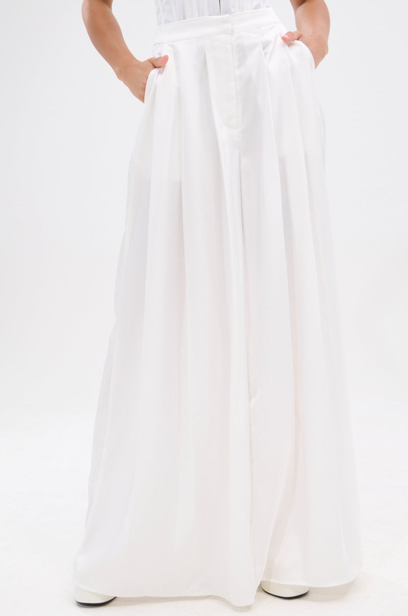 A person wearing the KYLA TROUSER, a high-rise, long, flowing white skirt with hands in the pockets, is standing against a plain white background. Only the lower body is visible; the upper body and head are not shown. 2