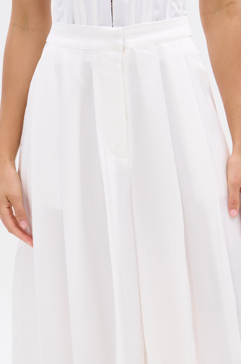 A person models the KYLA TROUSER, a white high-waisted, pleated pant with a chic high-rise silhouette, shown from waist to mid-thigh with arms relaxed at their sides. 6