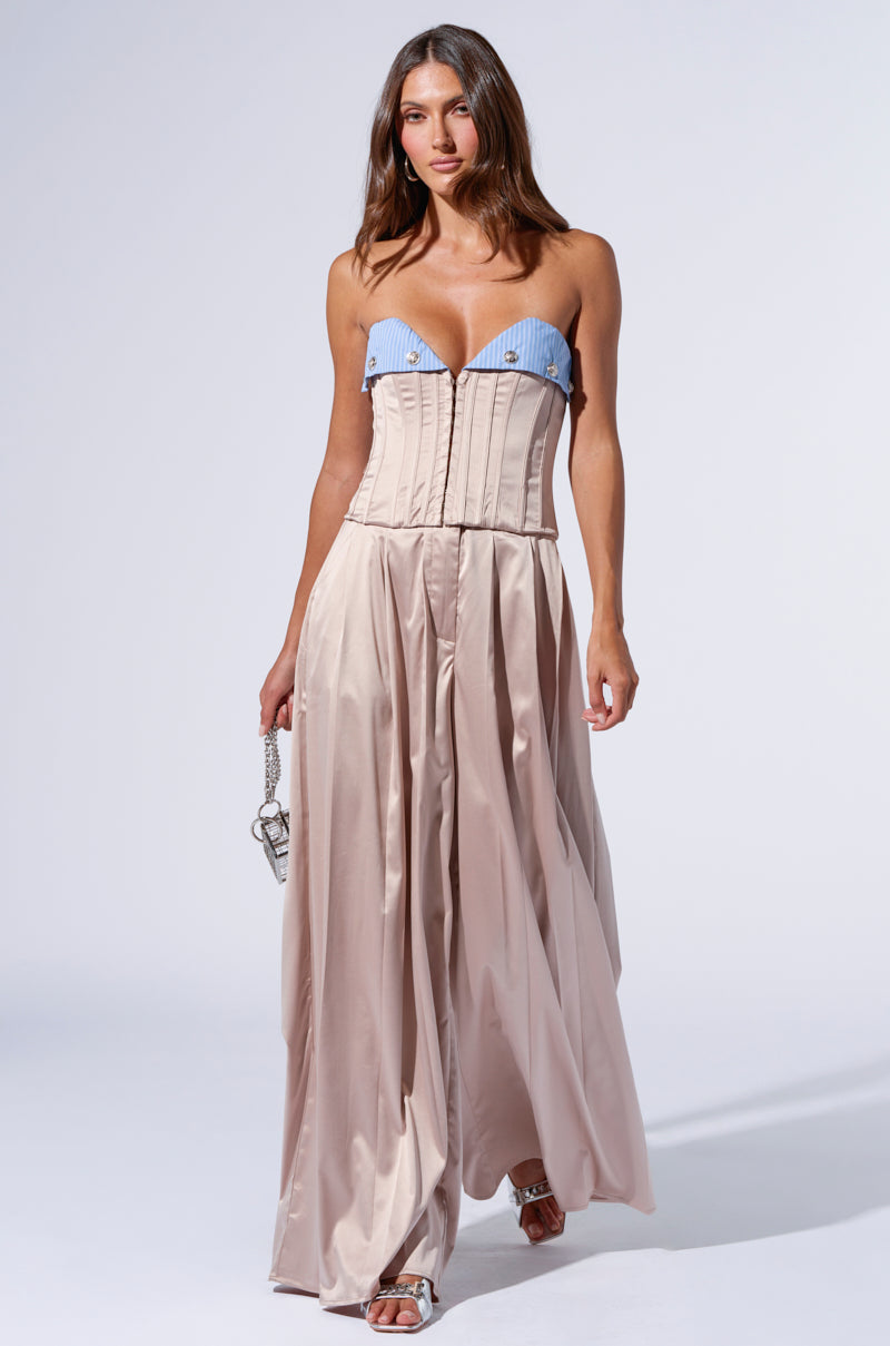 A woman is wearing the TAKE IT EASY CORSET TOP with a blue and white pattern and a long, flowing beige skirt. She holds a small handbag and wears silver high-heeled sandals. She has long, wavy hair and is walking forward on a plain, light-colored background, her outfit boasting delicate satin fabrication.