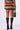 A person wearing the DRIVE ME WILD FUR MINI SKIRT, featuring layers of multicolored ombre faux fur, pairs it with high-heeled black leather boots and stands with their back to the camera. They hold a black bag embellished with metallic eyelets.