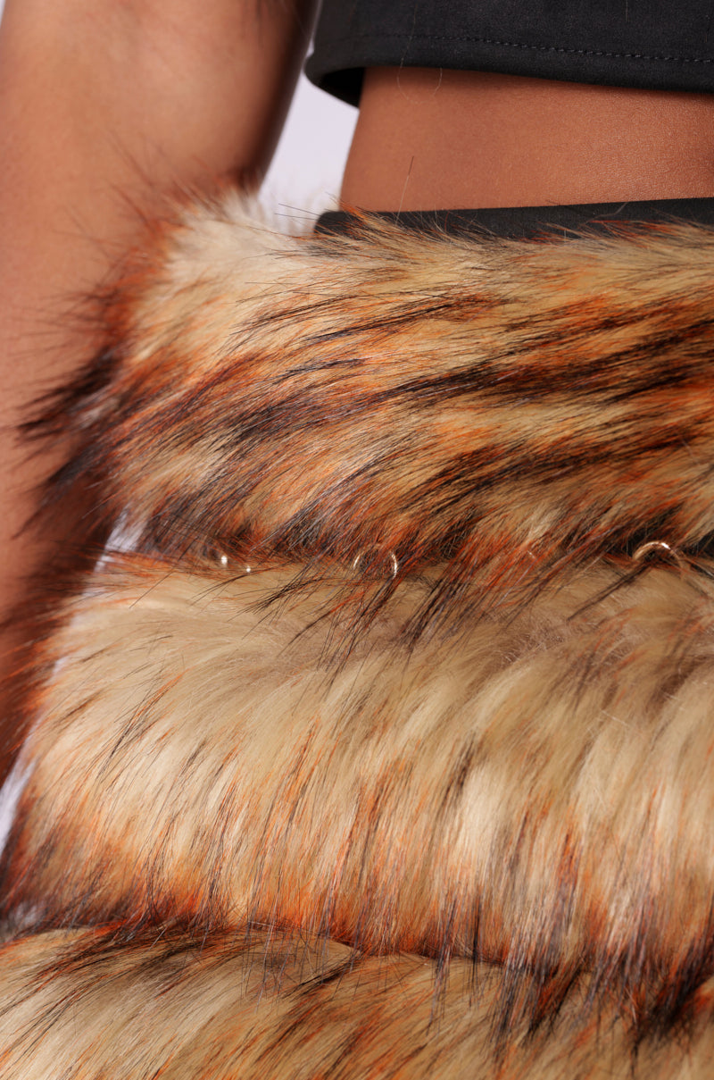 Close-up of a person's torso wearing a black top and the DRIVE ME WILD FUR MINI SKIRT, which features an ombre faux fur design with brown, beige, and orange streaks. The texture of the skirt is detailed and layered, creating a rich, voluminous appearance. The person's skin is visible at the top of the image.