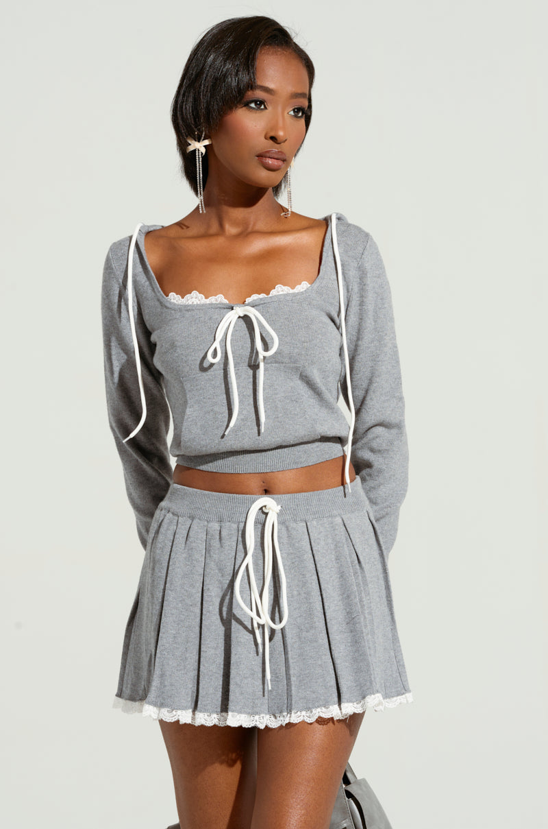 A woman poses in the PRETTY GIRL PROBLEMS TOP, a gray two-piece outfit with white lace trim that exudes a coquette vibe. This ensemble includes a cropped sweater accented with bow ties and is paired with a pleated skirt. With her short black hair and bold earrings, she stands against a plain background.