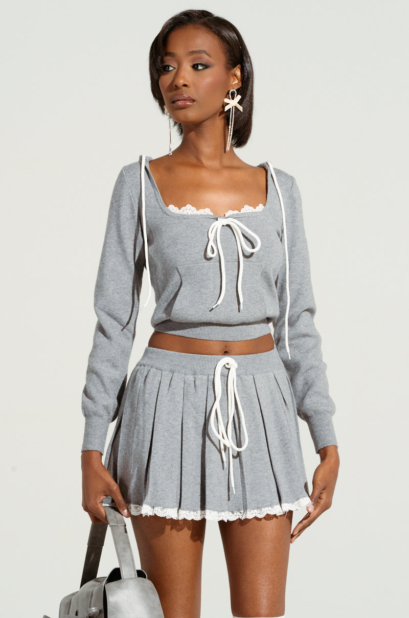 Dressed in the PRETTY GIRL PROBLEMS TOP, a person with long hair channels a coquette aura wearing a matching gray ensemble complete with a pleated skirt, both embellished with white lace and bows. They accessorize perfectly with dangling earrings while holding a gray bag against a plain background.