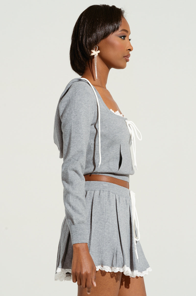 A person in profile is wearing the PRETTY GIRL PROBLEMS TOP, a gray cropped sweater paired with a matching pleated skirt accented by white lace trim, giving off a coquette vibe. They have long earrings and are standing against a plain background.