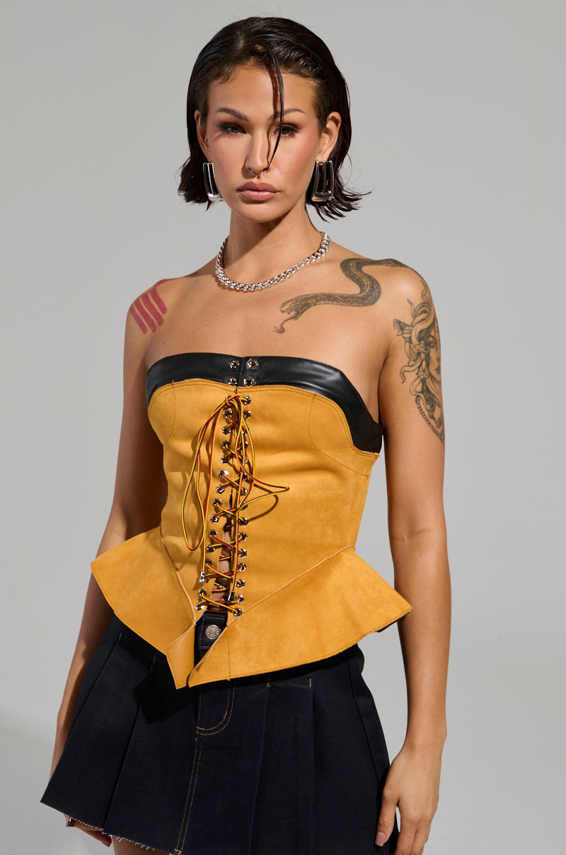 A person with short hair is wearing the NEW YORKS FINEST CORSET, a strapless yellow top with black trim and a lace-up front, paired perfectly with a dark pleated skirt. They showcase tattoos on their arms and shoulder, adding to their style with a necklace and hoop earrings to enhance the chic peplum silhouette.