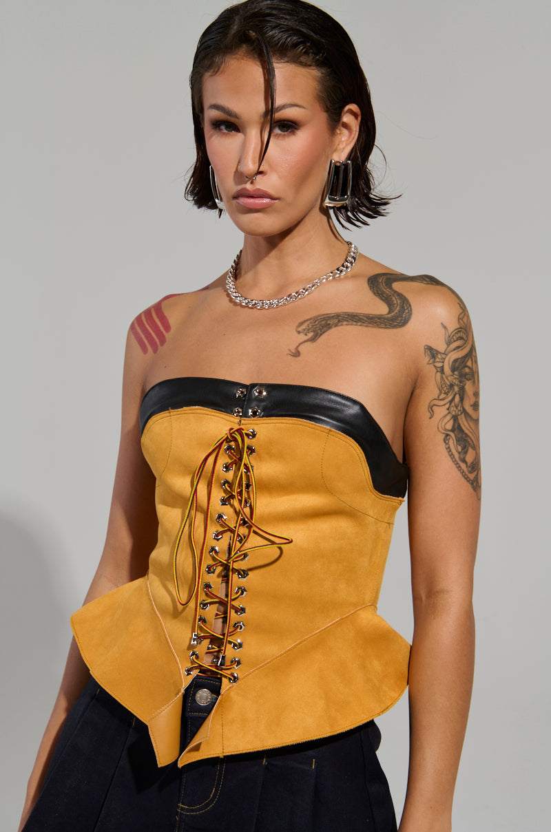 A person with short hair wears the NEW YORKS FINEST CORSET, a strapless yellow top accented with black leather trim and lacing. They showcase tattoos on both arms, paired with a silver chain necklace and square earrings, set against a plain gray backdrop.