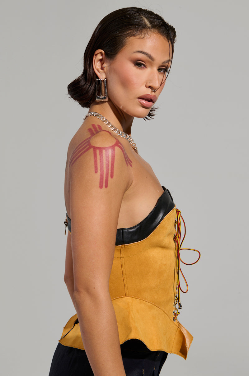 A woman with short dark hair showcases the NEW YORKS FINEST CORSET, a strapless yellow top accentuated with black trim and red lacing. Her look is completed with a red paint design on her shoulder, along with earrings and a chain necklace. The ensemble's subtle peplum silhouette adds an elegant touch as she gazes over her shoulder with a neutral expression.