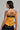 A person with tattoos on their arm and back is wearing the NEW YORKS FINEST CORSET, a strapless mustard-colored top with black trim and a back zipper. They have short dark hair, large hoop earrings, and are standing against a plain gray background.