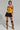 Person posing in a fashion-forward outfit, featuring the NEW YORKS FINEST CORSET in yellow, paired with a black mini skirt and faux suede tan knee-high lace-up boots. Tattoos are visible on their arms and thigh as they stand confidently against a neutral background.