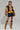 A person stands confidently in a faux fur jacket over the NEW YORKS FINEST CORSET in mustard yellow and a black pleated skirt. They wear knee-high tan lace-up boots and flaunt a tattoo on their thigh, all set against a plain gray background.