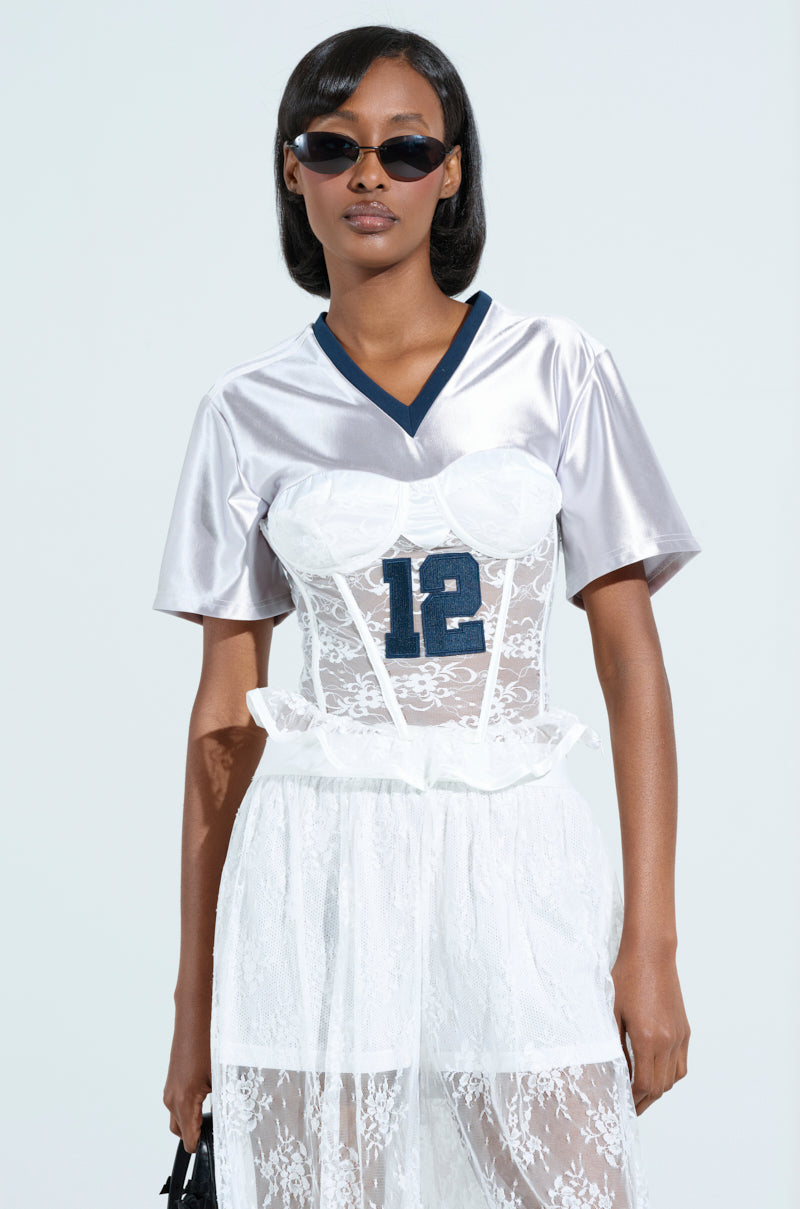 Wearing a LUCKY ME JERSEY with sporty accent stripes over a white lace corset and skirt, their short hair pairs well with dark sunglasses for a stylish look against the plain background.