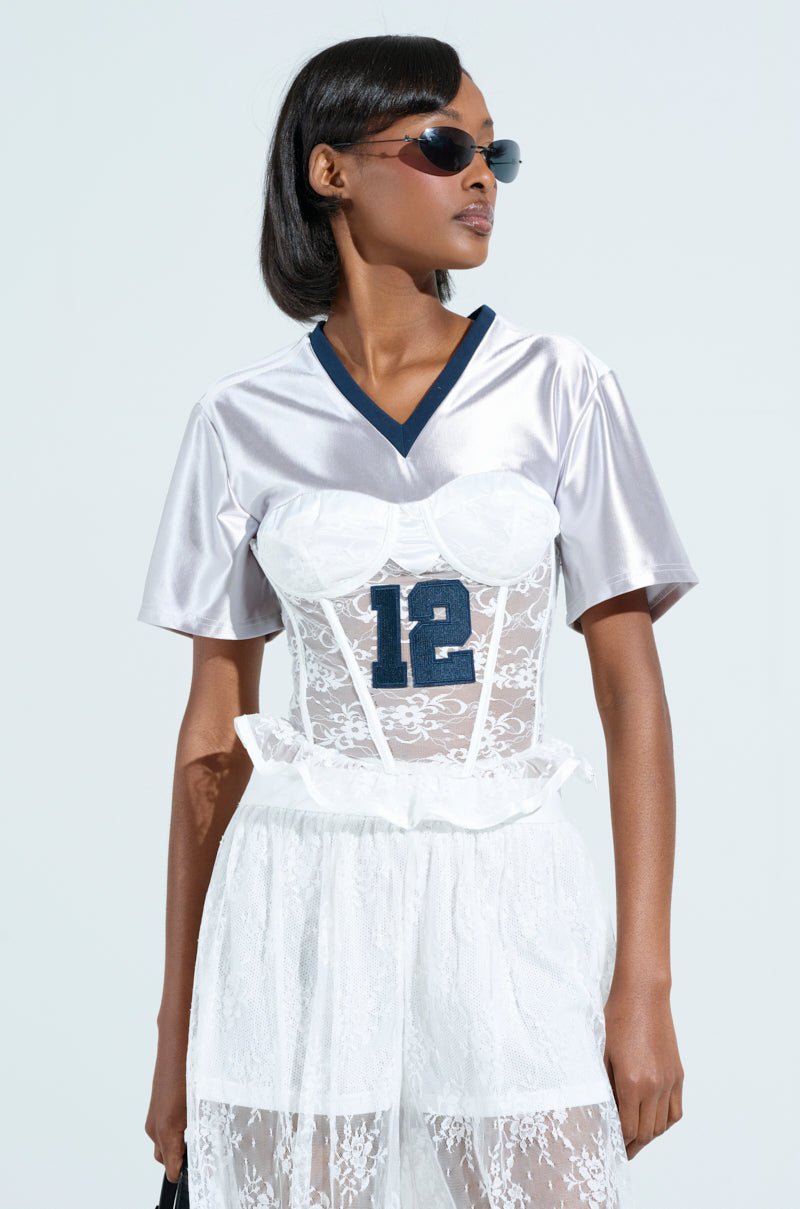 A stylish person exudes confidence with dark sunglasses and a chic short black bob, sporting a satin LUCKY ME JERSEY with a dark blue "12" and a white lace corset overlay, featuring sporty accent stripes against the plain background.