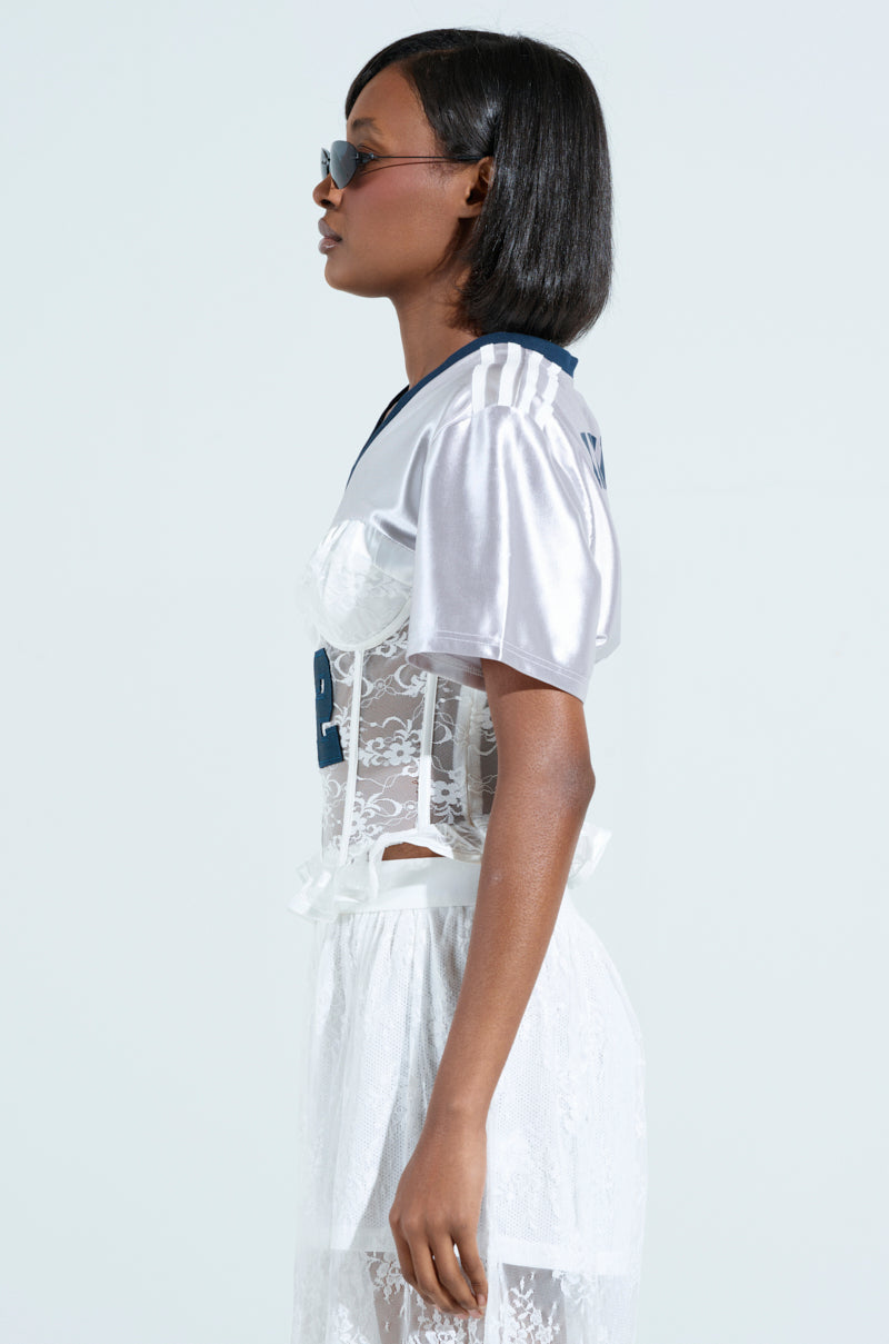 A model with a short bob and sunglasses wears a shiny LUCKY ME JERSEY with the number 22. The jersey is layered over sheer lace adorned with ribbons and features a corseted lower-half against a light background.