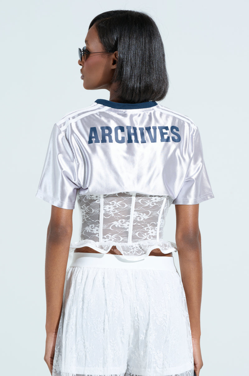 A person with shoulder-length black hair and sunglasses wears a shiny silver LUCKY ME JERSEY over a lace corset, paired with a white lace skirt featuring sporty accent stripes. The backdrop is plain light blue.