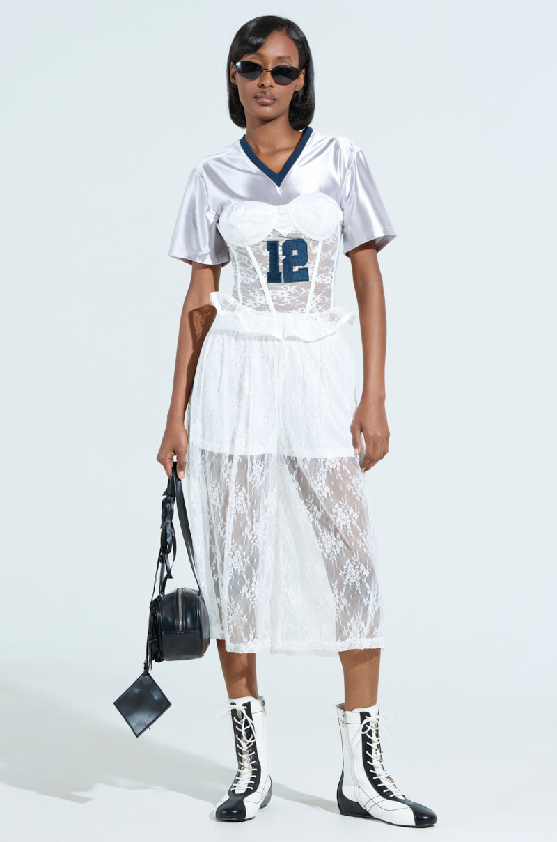 Wearing a LUCKY ME JERSEY with sporty accent stripes under a white lace dress, paired with sunglasses, black and white boots, and a black round bag against a light gray backdrop, creates an ensemble of bold style.