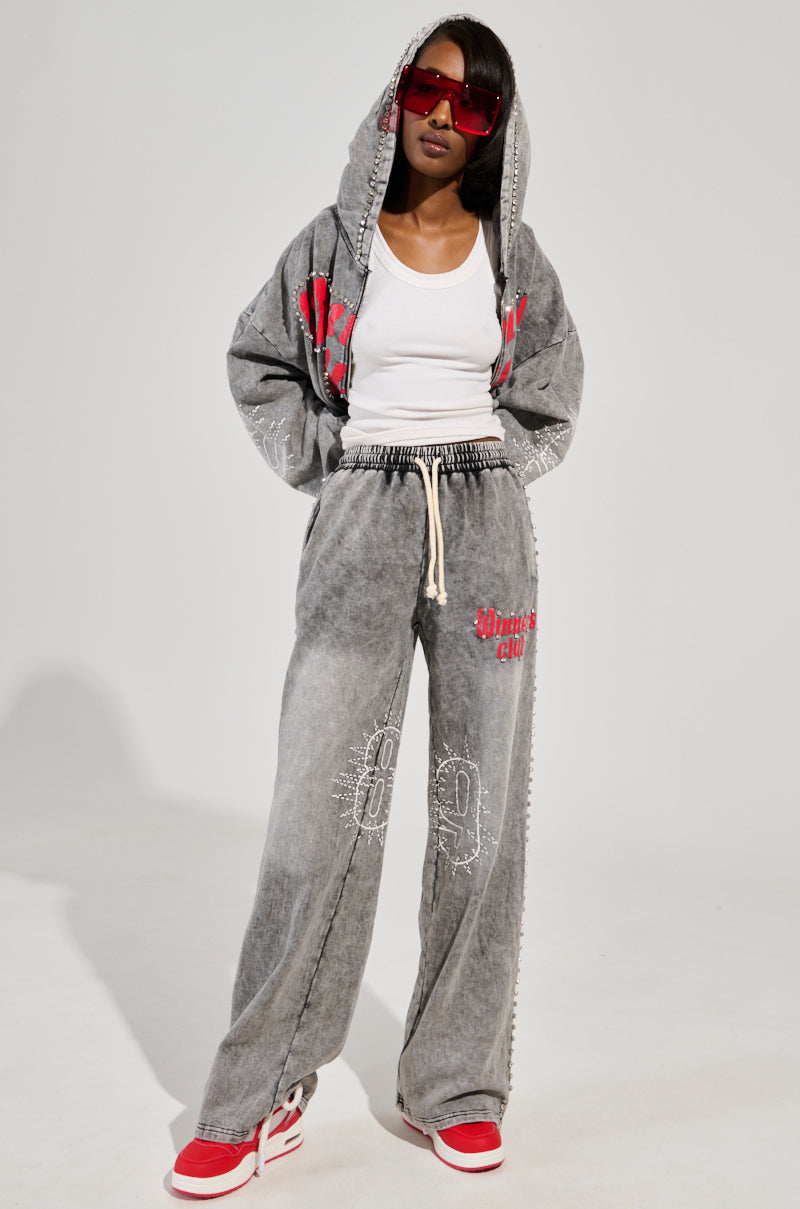 Wearing a white tank top, the WINNERS CLUB ZIP UP SWEATSHIRT, and matching wide-leg pants with red accents from the AKIRA Label Winners Club collection, along with oversized red sunglasses and sneakers, a person poses confidently against a plain background in chic loungewear.