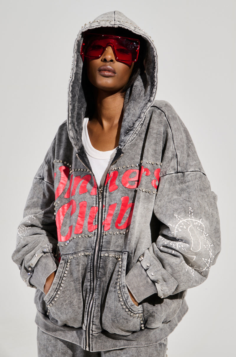 A person confidently wears a WINNERS CLUB ZIP UP SWEATSHIRT, a gray studded hoodie with a red graphic from the AKIRA Label collection, paired with matching pants. They're styled with oversized red sunglasses and a white undershirt, embodying an oversized loungewear look with modern flair.