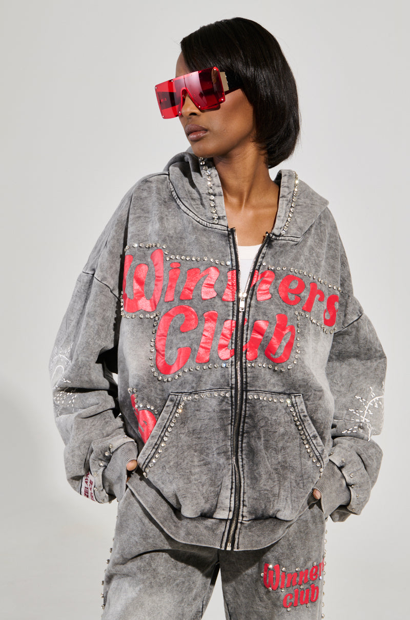Sporting oversized loungewear, a person wears a grey AKIRA Label Winners Club hoodie and pants with red "Winners Club" text. They pair it with large red sunglasses, and the outfit resembles a studded rhinestone sweatshirt, as they stand casually with hands in pockets.