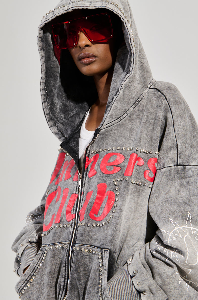 A person is wearing an oversized gray WINNERS CLUB ZIP UP SWEATSHIRT featuring "Dreamers Club" in bold red. Large red sunglasses complement the look over a crisp white shirt, with rhinestones adding sparkle to the sleeves.