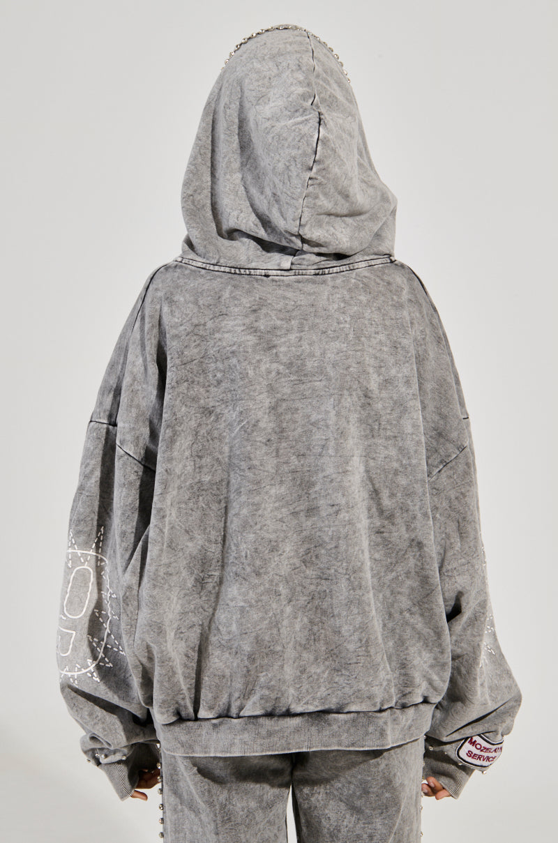 Someone is wearing an oversized gray AKIRA Label Winners Club Zip Up Sweatshirt with matching pants, featuring subtle white designs on the sleeves and back. The hood is pulled up, obscuring their head against a plain light background.