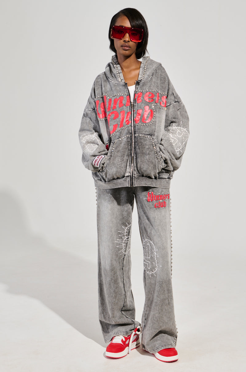 Wearing oversized dark sunglasses and the AKIRA Label Winners Club Zip Up Sweatshirt, a person pairs it with matching baggy pants showcasing red text and designs. They stand confidently on a plain background, completing the look with red and white sneakers, hands casually in pockets.