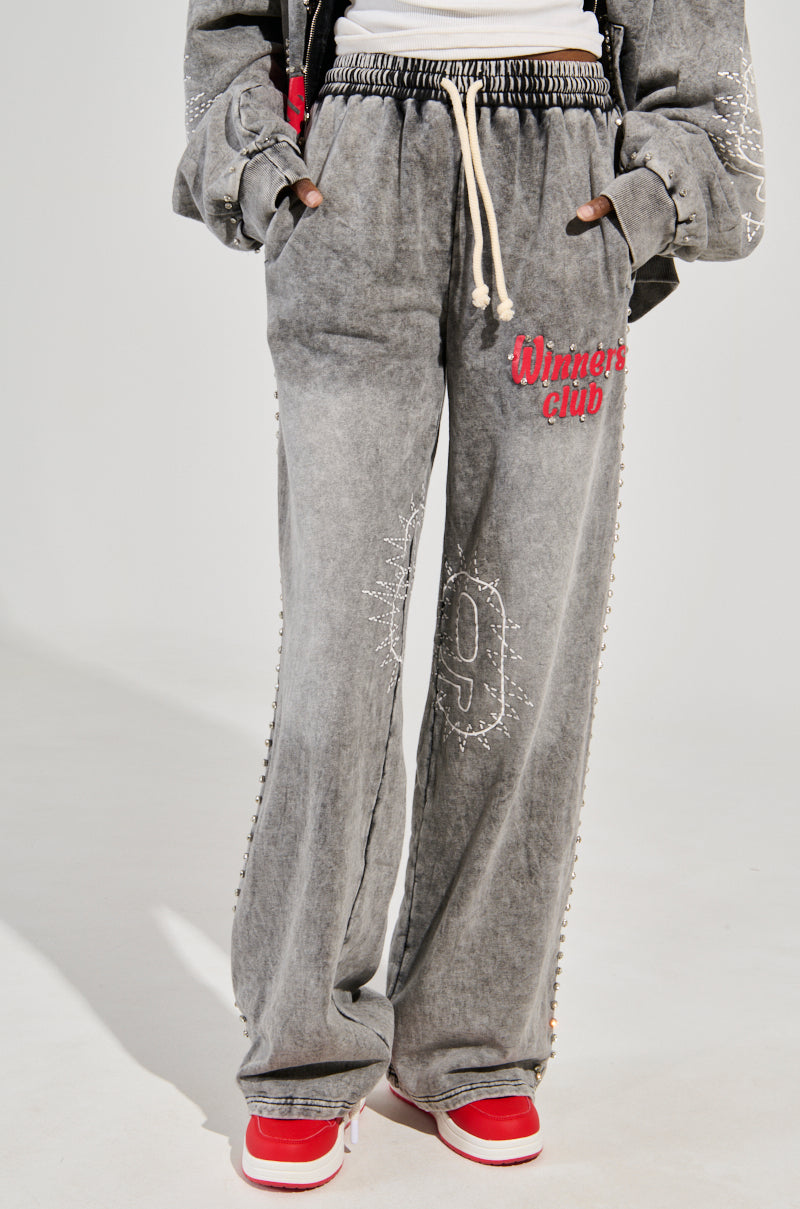 A person stands confidently in their gray WINNERS CLUB WIDE LEG JOGGER, featuring "Winners Club" in red, studded accents, and a bold number "9." With hands in pockets, they pair it with stylish red and white sneakers.