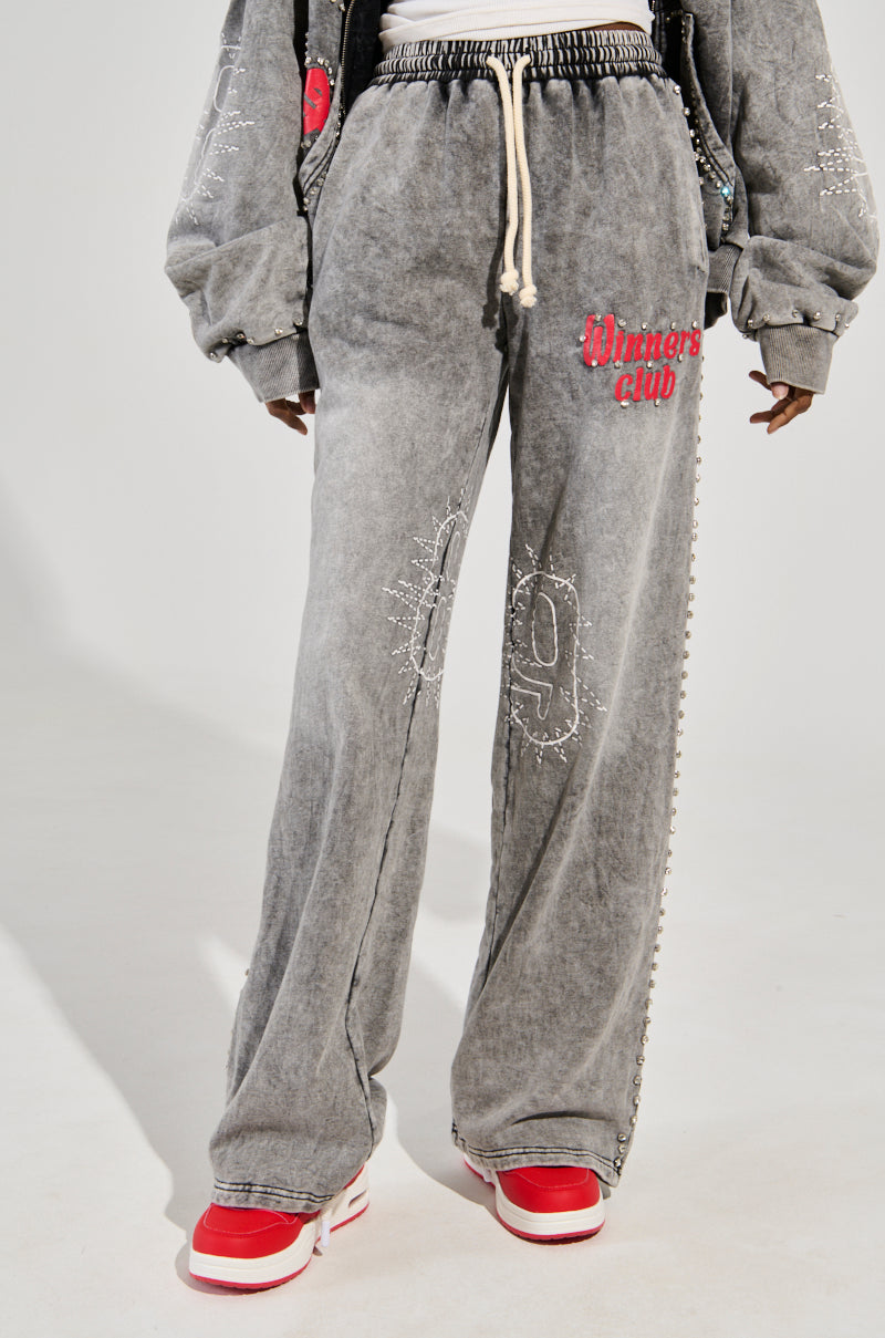 Wearing faded gray WINNERS CLUB WIDE LEG JOGGER sweatpants with red embroidered details and a drawstring waist, paired with red and white sneakers, creates a stylish yet laid-back look.