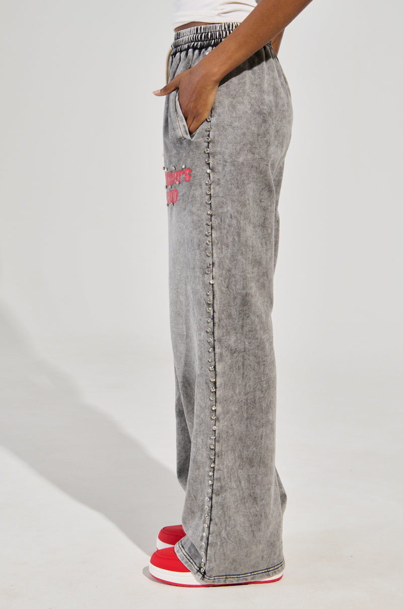 A person models the WINNERS CLUB WIDE LEG JOGGER sideways in gray acid-wash, featuring a drawstring waist and bold red "Winners Club" text on the side. They're styled with red and white sneakers for a casual look against a simple white background.