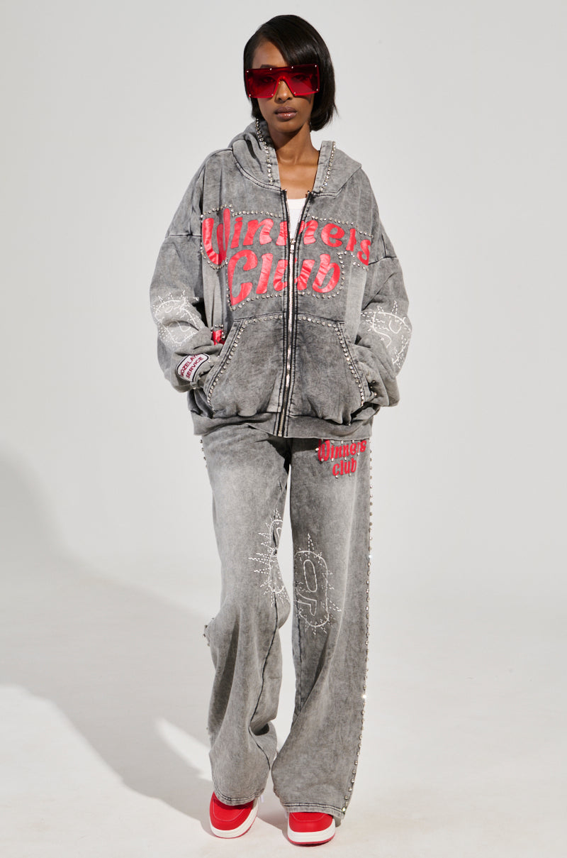 A person confidently wears an oversized gray cotton knit hoodie and WINNERS CLUB WIDE LEG JOGGER, both featuring red text, paired with red sneakers. They sport large red sunglasses against a plain backdrop.