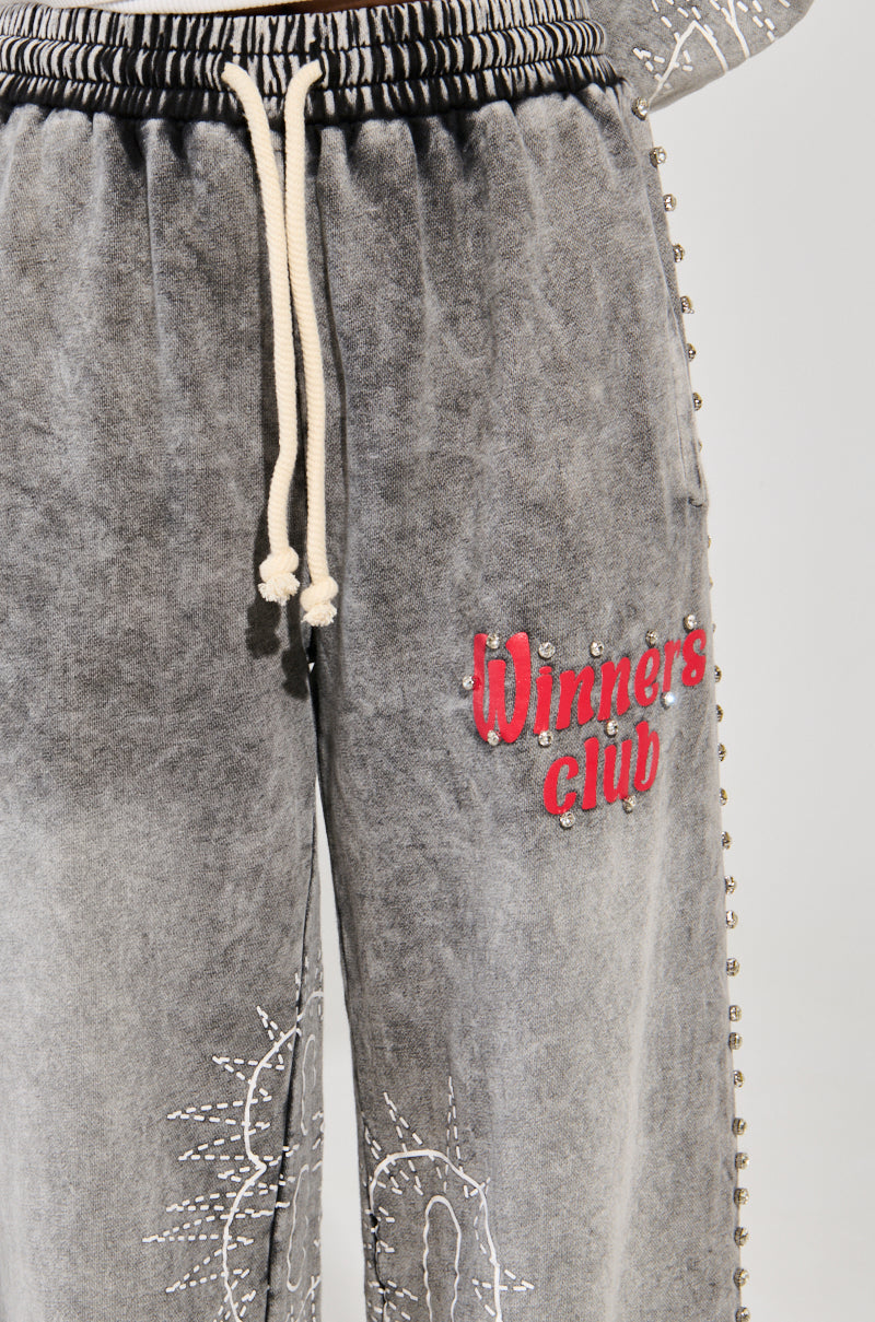 Close-up of gray WINNERS CLUB WIDE LEG JOGGER sweatpants, featuring a drawstring waist, stud embellishments along the sides, and "Winners Club" embroidered in red on the leg. The cotton knit fabric provides a weathered, vintage look.