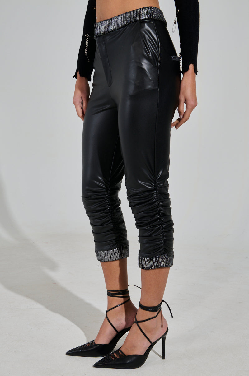 A person is wearing the Always Best Dressed Capri in black PU, featuring shiny faux leather with ruched legs and metallic cuffs. The high-rise design includes zipper details and is paired with black lace-up heels and a black long-sleeve top. 2