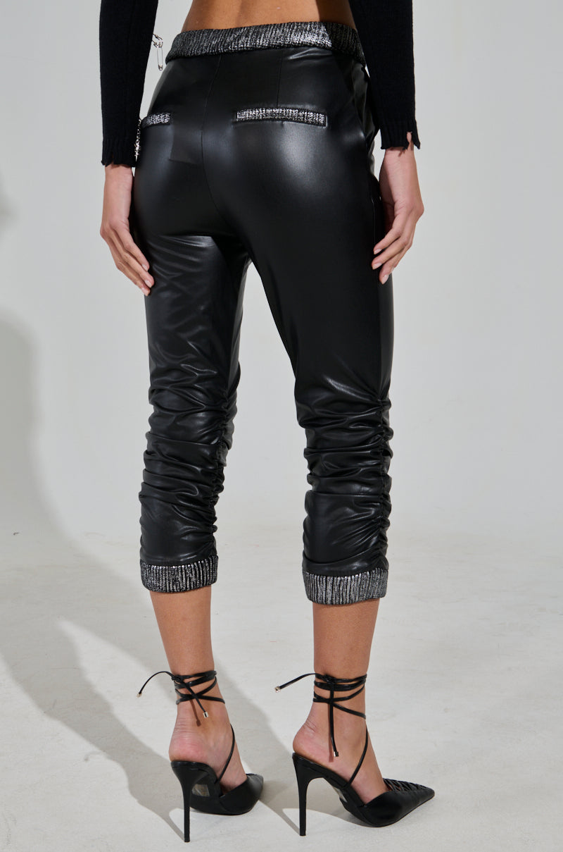 A person is wearing ALWAYS BEST DRESSED CAPRI IN BLACK PU, featuring black faux leather jogger pants with sparkly cuffs, a matching waistband, and zipper details. They are paired with black high heels with ankle straps on a light background. 3