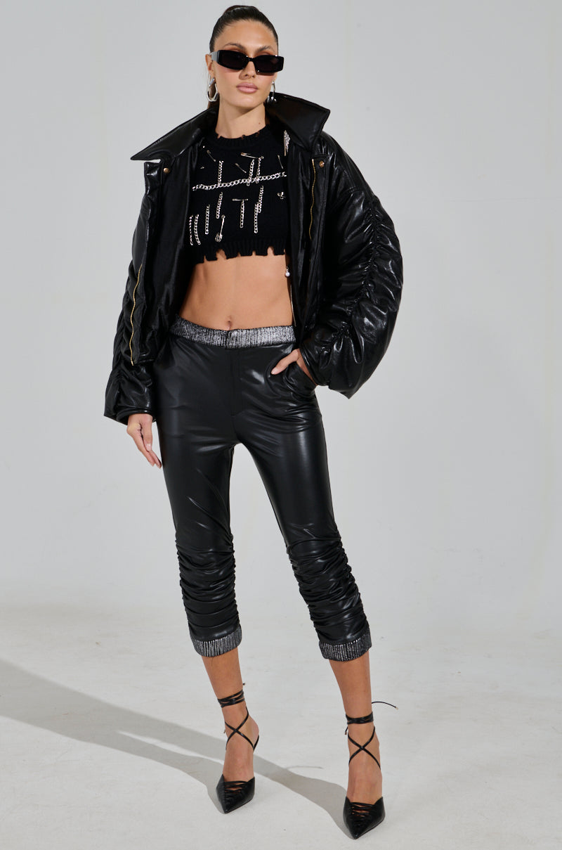 A person confidently dons a shiny black jacket, a black cropped top with a cross design, and the ALWAYS BEST DRESSED CAPRI IN BLACK PU high-rise faux leather pants, complemented by black heels, long hair tied back, and dark sunglasses. 4