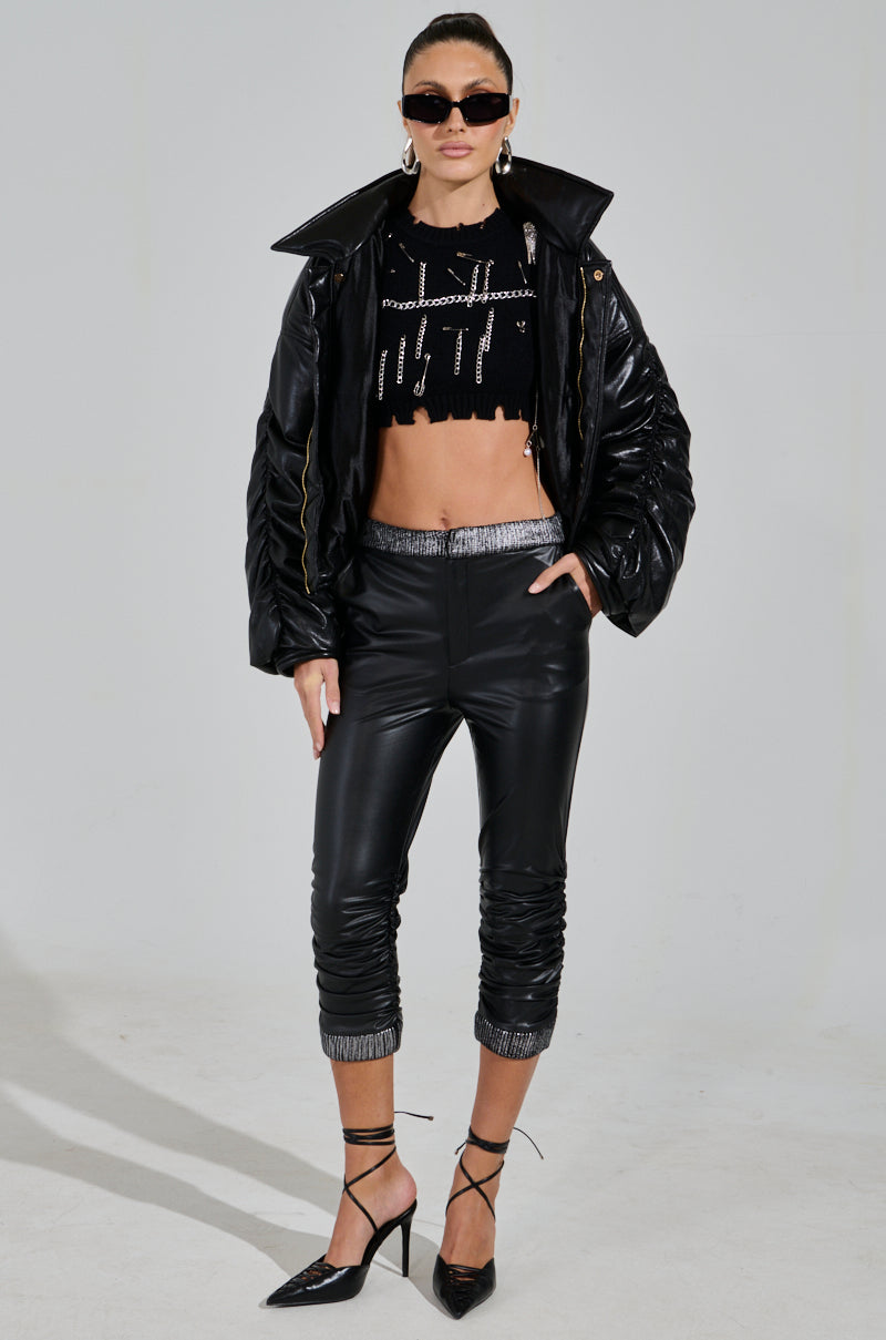 Against a gray background stands a person in the ALWAYS BEST DRESSED CAPRI IN BLACK PU, a cropped top, and a black faux leather jacket. With sunglasses and high heels on, they exude confidence, their hands resting in their jacket pockets. 5