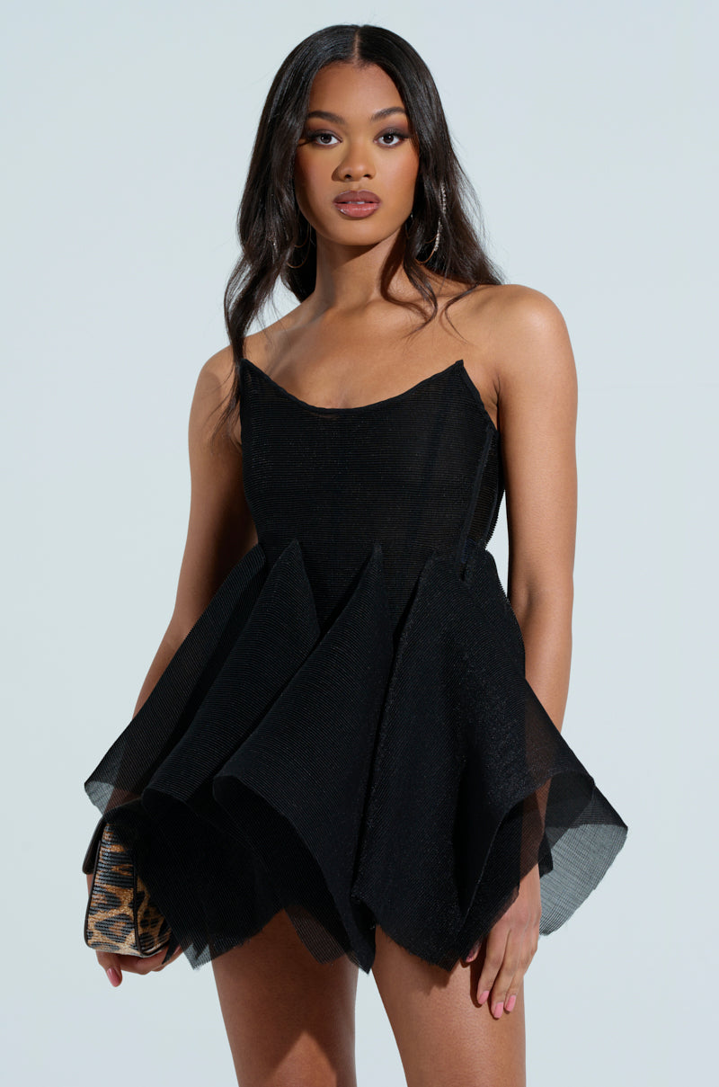 A woman confidently poses against a plain background, wearing a CRAZY 4 YOU BLOUSE—a stylish, short black dress with a flared skirt and sheer mesh top. She holds a small leopard print purse and has long, wavy hair.