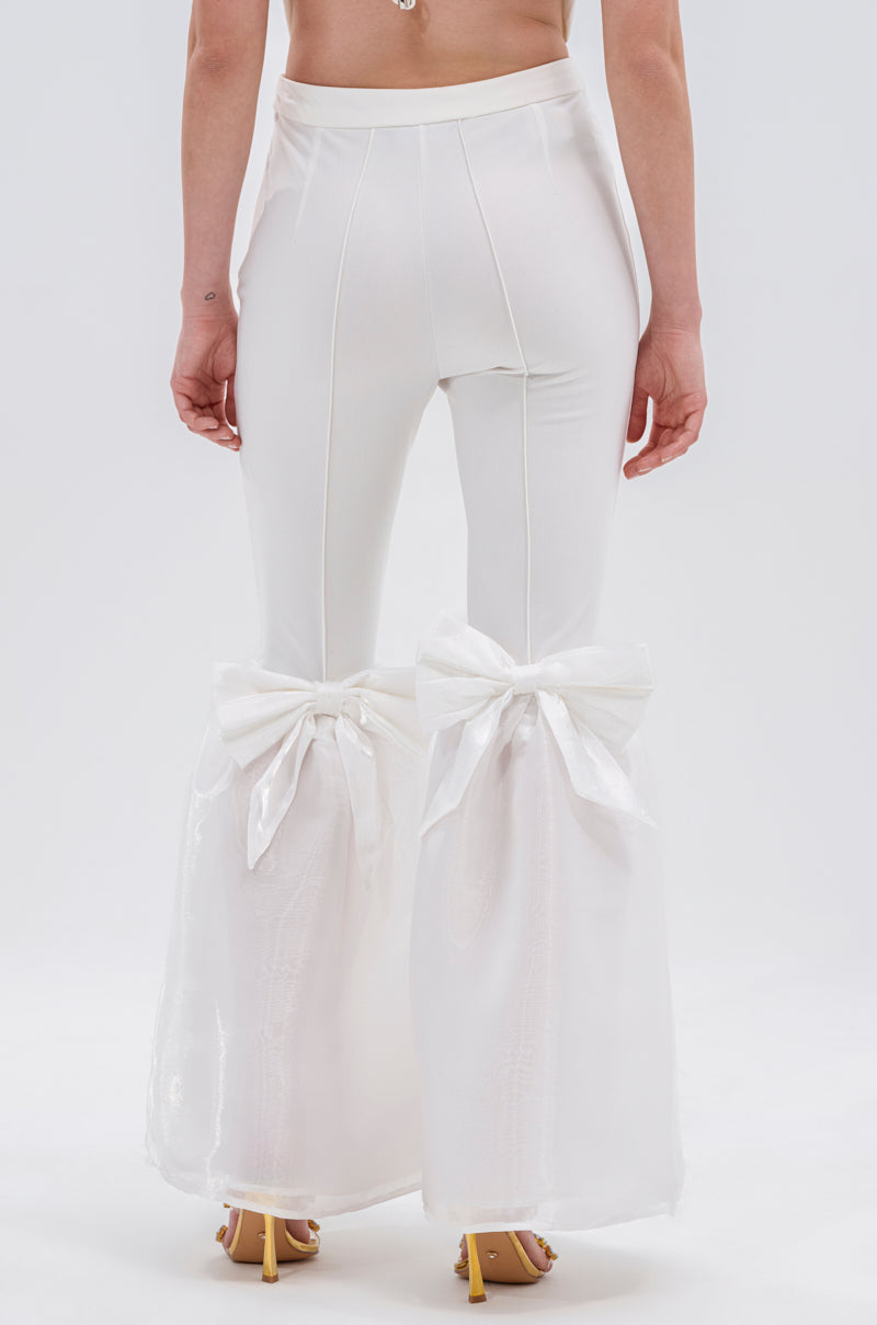 A person wears the LILY TROUSER WITH BOW—high-waisted, white flared pants with sheer fabric and satin bows at the back—paired with open-toe heels. The photo is taken from behind against a plain background.