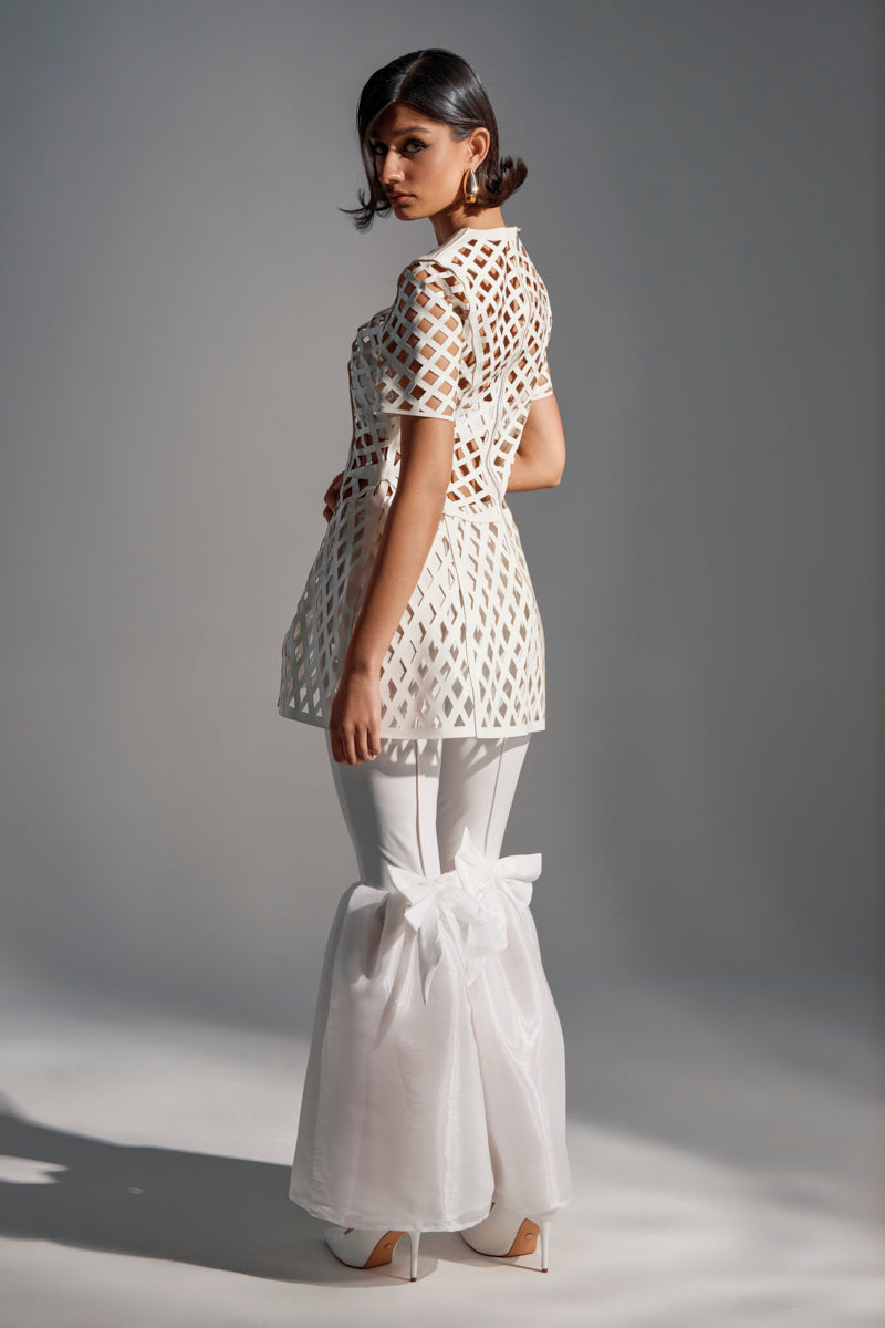 A woman poses in a geometric white short-sleeve top and the LILY TROUSER WITH BOW, featuring high-rise sheer wide flares tied at the knees with satin bows, set against a softly lit plain background.