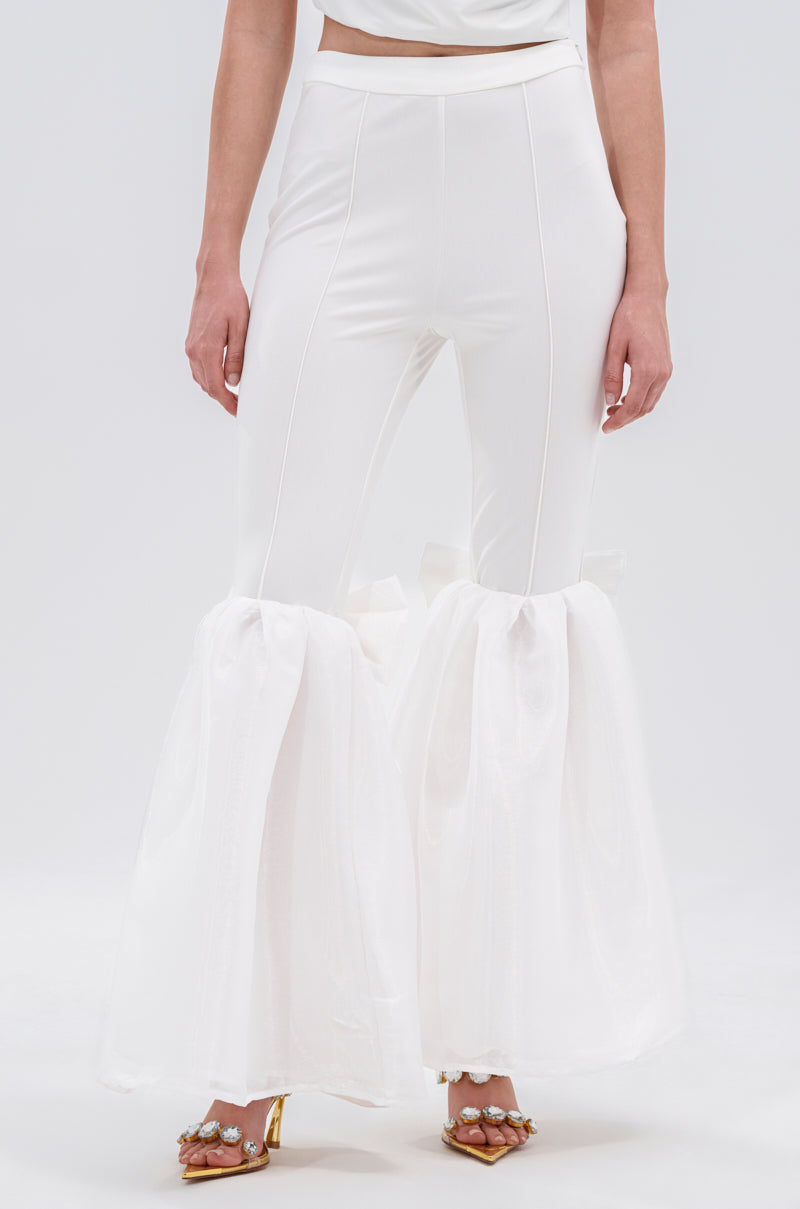 A person wears white LILY TROUSER WITH BOW, featuring a high-rise waist and sheer ruffles at the lower legs, paired with gold embellished heels. The cropped photo shows only the lower body against a plain light background.