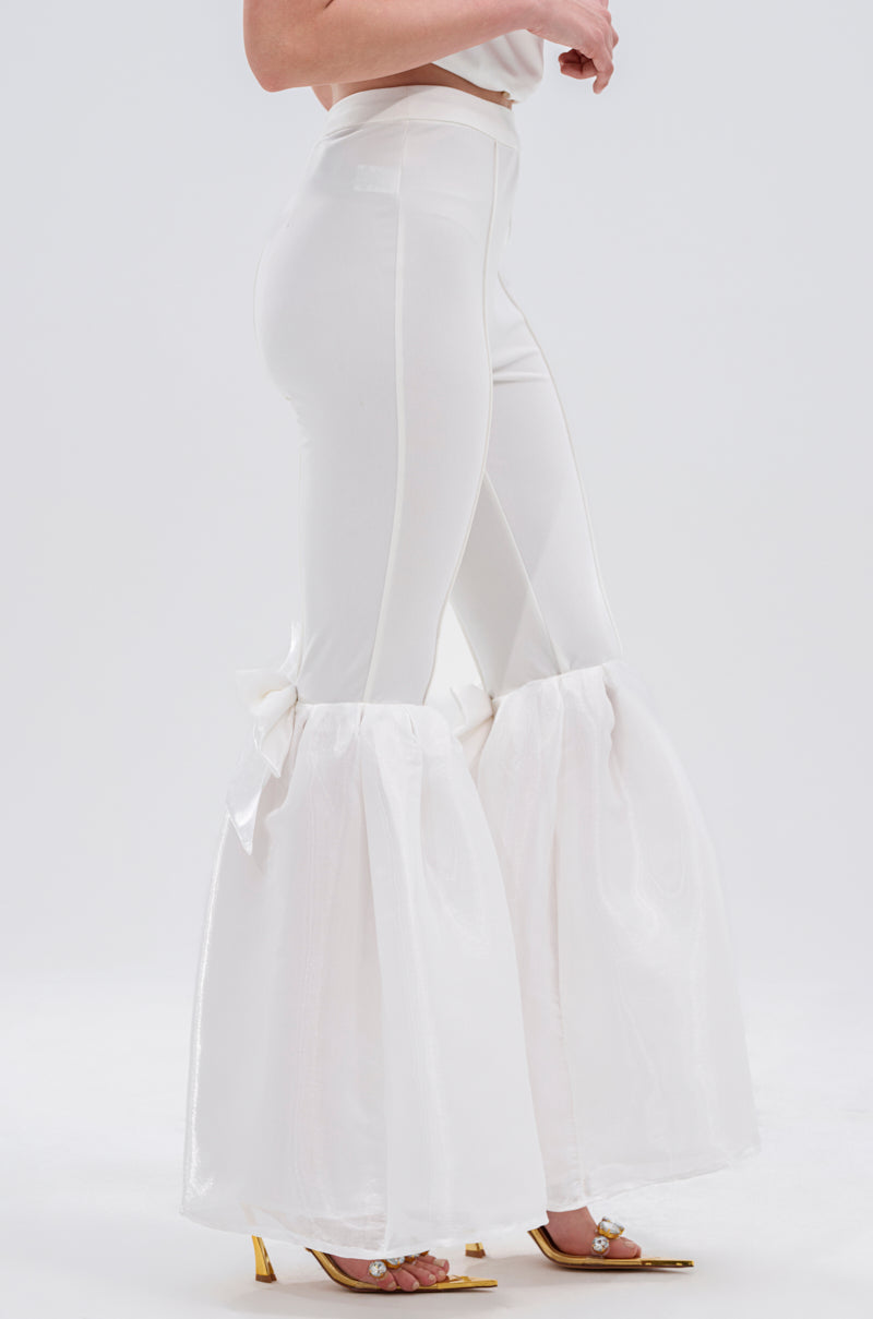 A person models the LILY TROUSER WITH BOW, featuring a high-rise waist and sheer, flared bottoms, styled with gold open-toe heels against a plain white background.