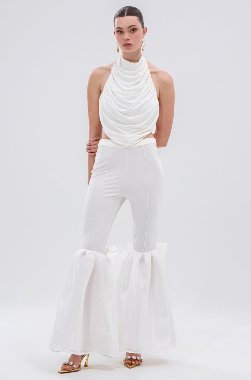 A woman with slicked-back hair wears gold earrings, gold open-toe heels, a white sleeveless draped top, and the LILY TROUSER WITH BOW—high-rise elegant trousers featuring sheer flared ruffles at the calves—against a plain background.