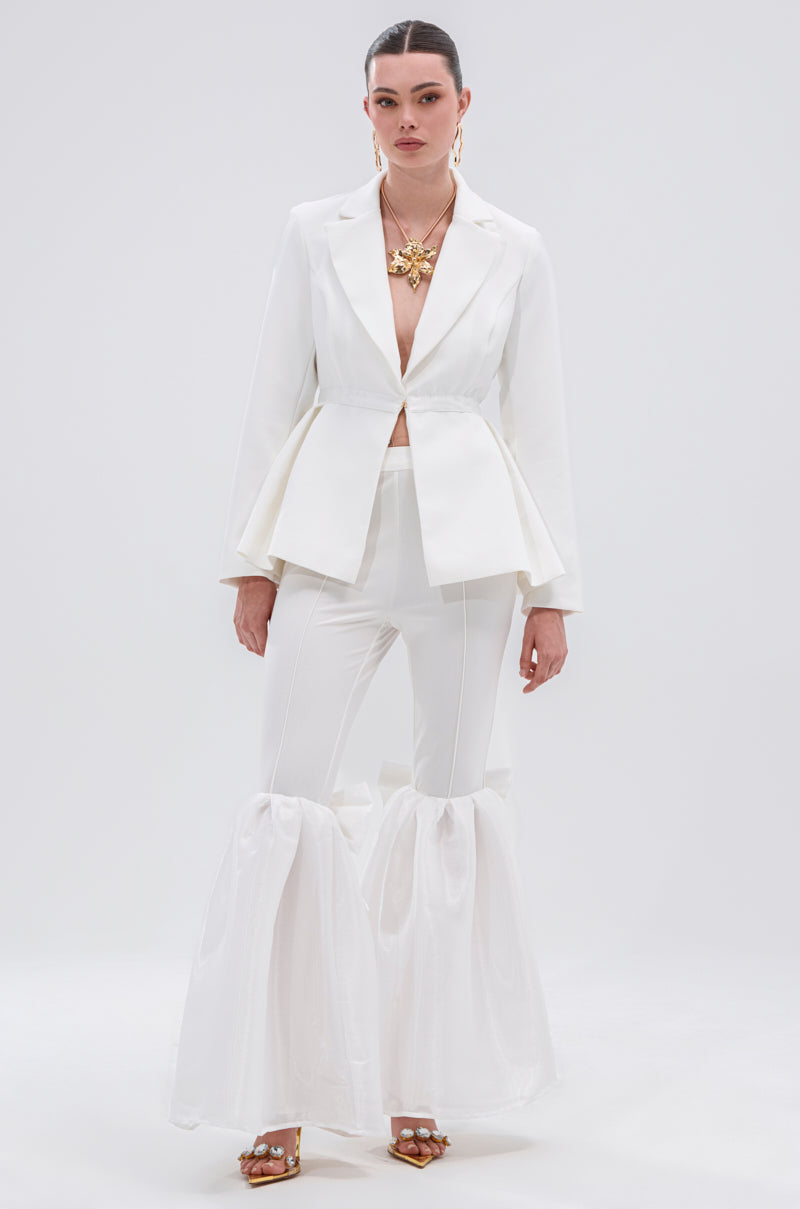 A woman stands against a plain background wearing a tailored white blazer, Lily Trouser with Bow featuring a high-rise waist and sheer, voluminous ruffles at the ankles, gold jewelry, and open-toed heels with gold details.