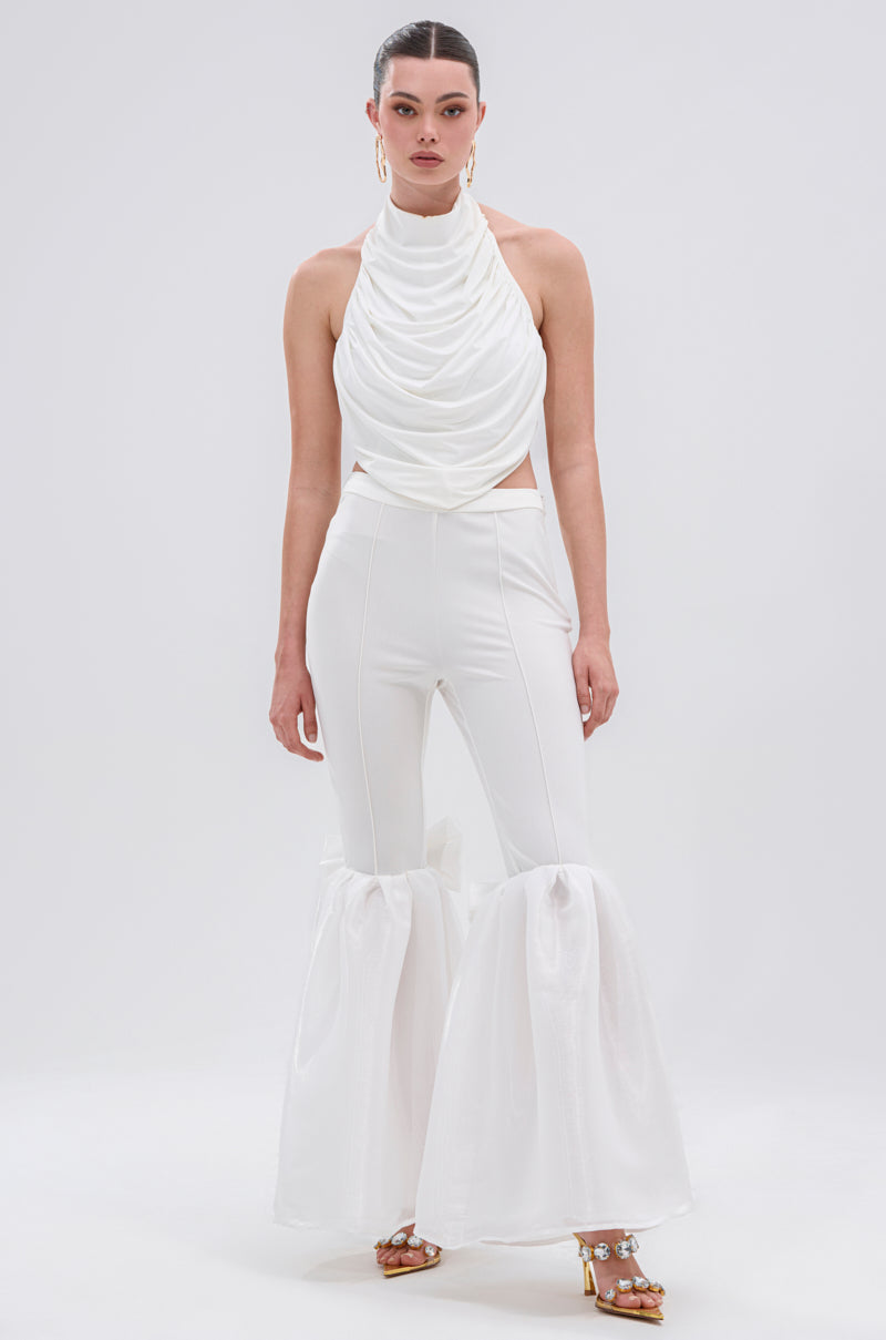 A woman with slicked-back hair and gold hoop earrings wears an all-white outfit featuring a draped halter top and the LILY TROUSER WITH BOW, which has a high-rise waist and dramatic sheer ruffles at the knees.